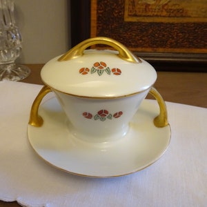 May include: A white porcelain creamer with a gold trim and red and green floral design. The creamer has two gold handles and a lid with a gold knob.