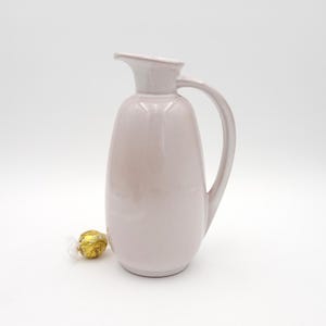 May include: A pale pink ceramic jug with a curved handle and a spout. The jug has a smooth, glossy finish and a rounded body. A wrapped gold sweet sits near the base of the jug.
