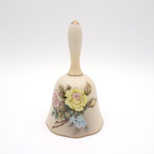 May include: Cream-colored ceramic bell with a long handle and a floral design. The bell features a bouquet of yellow, pink, and blue flowers with green leaves. The handle has a gold-colored tip.