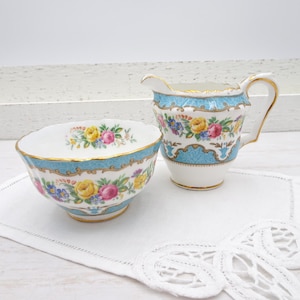 Op de afbeelding: Een vintage porseleinen melk- en suikerpotset. De set heeft een witte basis met een lichtblauwe rand en gouden afwerking. Bloempatronen in geel, roze en blauw sieren de stukken. De melkkan heeft een handvat en een tuit.