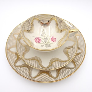 May include: A vintage teacup, saucer, and dessert plate set. The white porcelain set features a gold honeycomb pattern and scalloped edges. The teacup and plate have a floral design with pink flowers and gray leaves.