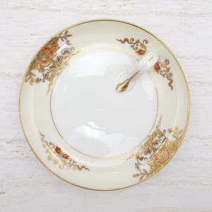 May include: A round, off-white ceramic dish with a gold rim and handle. The dish features floral and scroll designs in gold, red, and blue. The handle is gold-colored. The dish is likely for serving or display.