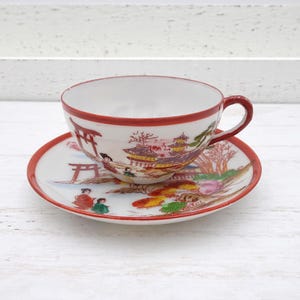 Op de afbeelding: Een wit theekopje en schotel met een rode rand en een Japans geïnspireerd ontwerp met een rode poort, gebouwen en figuren in een landschap.