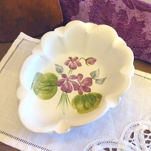 Puede incluir: Un plato de cerámica blanca con borde festoneado presenta un diseño pintado a mano de violetas moradas y hojas verdes.