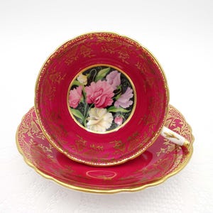 Puede incluir: Una taza de té y un platillo rosas con un borde dorado y un diseño floral. La taza presenta un fondo negro con un ramo de guisantes de olor rosas, blancos y morados.