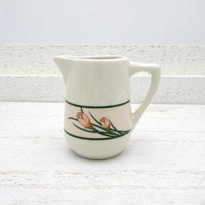 May include: A small, off-white ceramic pitcher with a handle and spout. A pink band with orange floral designs and green accents encircles the pitcher. The pitcher is likely used for serving cream or other liquids.
