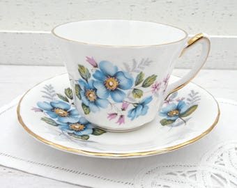 May include: A white teacup and saucer with a blue floral pattern. The cup and saucer have a gold trim.
