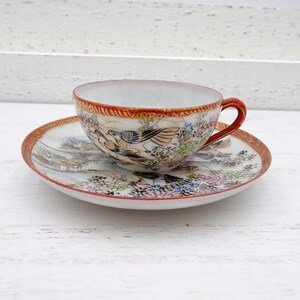 May include: A vintage teacup and saucer set. The cup and saucer are white with a detailed painted design featuring birds, flowers, and a landscape scene. The rims are trimmed with a red and gold border. The cup has a red handle.