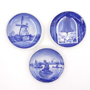 Puede incluir: Tres platos pequeños de cerámica azul y blanca con diferentes escenas holandesas y danesas. Los platos muestran un molino de viento, un campanario y una sirena.