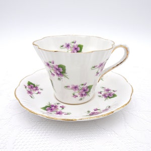 May include: A white teacup and saucer with a delicate floral pattern of purple violets. The cup and saucer have a gold trim around the edges.