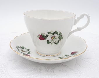 May include: A white porcelain teacup and saucer set featuring a delicate floral pattern of white blossoms, green leaves, and a single red bud. The teacup has a scalloped rim and a gold-trimmed edge, with a matching scalloped saucer.