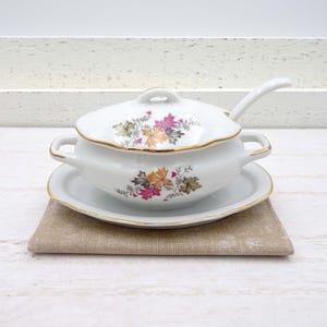 May include: White porcelain gravy boat with a lid and a matching saucer. The boat and saucer have a gold trim and a floral design with pink, orange, and green leaves.