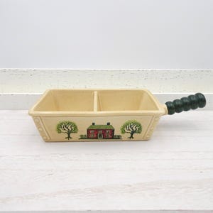 May include: Cream ceramic divided serving dish with a green wooden handle. The dish is decorated with a red house, green trees, and a green handle. The dish is approximately 30 cm long.