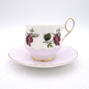Puede incluir: Un juego de taza y platillo de té blanco con un borde rosa y ribete dorado. La taza de té presenta un diseño floral con rosas rojas y hojas verdes.