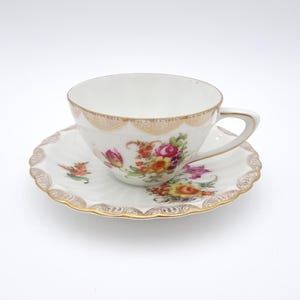 May include: A white porcelain teacup and saucer set with a floral pattern in shades of pink, yellow, and orange. The set has a gold trim and a scalloped edge.