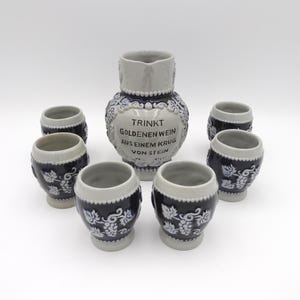 May include: A blue and white ceramic wine set, comprising a pitcher and six small mugs. The pitcher and mugs feature a grape vine design. The pitcher has the text "TRINKT GOLDENEN WEIN AUS EINEM KRUG VON STEIN".