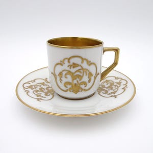 May include: A white porcelain teacup and saucer set featuring intricate gold filigree floral patterns. The cup has a gold-lined interior and a gold-painted handle, while the saucer features matching gold scrollwork along the outer rim.