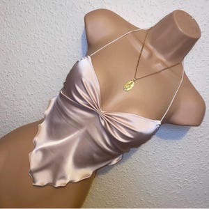 May include: A pale pink satin camisole top featuring a V-neckline with gathered fabric at the centre, thin spaghetti straps, and a pointed handkerchief hemline. A gold-toned oval pendant necklace rests against the fabric of the garment.