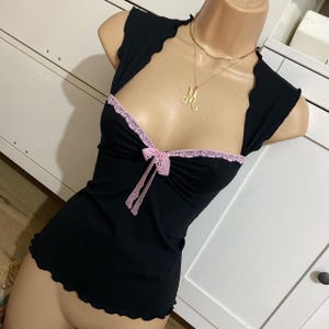 May include: Black sleeveless top with a scalloped neckline and hem. The top has a pink lace trim and a small bow. A gold necklace with the letter "M" is also visible.
