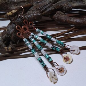 Puede incluir: Un par de aretes largos y colgantes con cuentas turquesa y blancas, alambre de cobre y detalles de concha blanca.