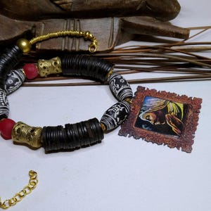 Peut inclure: Un collier artisanal avec des perles noires, dorées, rouges et blanches. Le collier comprend des perles noires cylindriques, des accents métalliques dorés et des perles blanches à motifs. Une petite image encadrée est également présente.