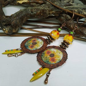 May include: A pair of dangle earrings featuring circular copper-toned textured frames with sunflower imagery. Each earring includes a yellow and orange patterned glass bead, small green and orange accent beads, and a yellow teardrop-shaped charm.