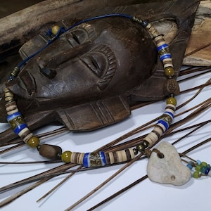 May include: Handmade necklace featuring a heart-shaped stone pendant. The necklace incorporates wooden beads, blue and white glass beads, and small green and blue beads. The necklace is strung on a blue cord and has a rustic, bohemian aesthetic.