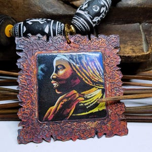 May include: A handcrafted necklace featuring a pendant with a portrait of a woman with yellow and gold braids. The pendant is framed in a decorative, reddish-brown border. The necklace includes black and white patterned beads and wooden beads.