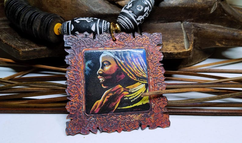 May include: A handcrafted necklace featuring a pendant with a portrait of a woman with yellow and gold braids. The pendant is framed in a decorative, reddish-brown border. The necklace includes black and white patterned beads and wooden beads.