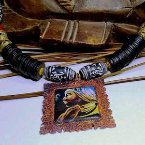 May include: Handmade necklace featuring black, white, and red beads with gold-colored accents. The necklace includes a pendant with a portrait of a person in a decorative frame. The necklace is displayed on a light surface.