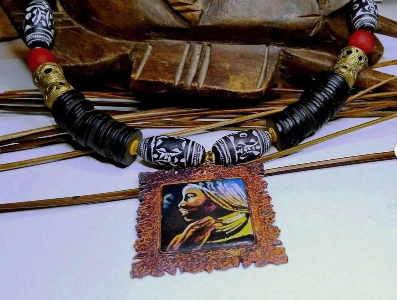 May include: Handmade necklace featuring black, white, and red beads with gold-colored accents. The necklace includes a pendant with a portrait of a person in a decorative frame. The necklace is displayed on a light surface.