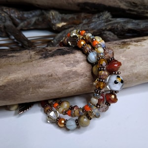 May include: A triple-strand bracelet featuring brown, orange, and white beads. The beads are made of glass and come in a variety of shapes and sizes. The bracelet is displayed on a wooden surface.