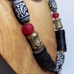 May include: A handcrafted necklace featuring black and white patterned beads, red glass beads, and gold-colored metal accents. The necklace is displayed on a wooden bust, showcasing its unique design and cultural aesthetic.