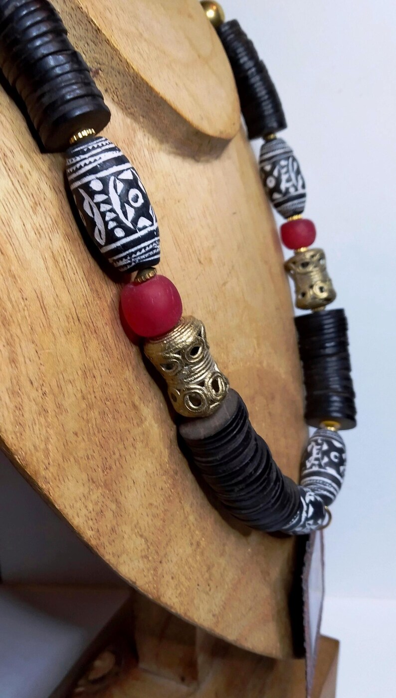 May include: A handcrafted necklace featuring black and white patterned beads, red glass beads, and gold-colored metal accents. The necklace is displayed on a wooden bust, showcasing its unique design and cultural aesthetic.