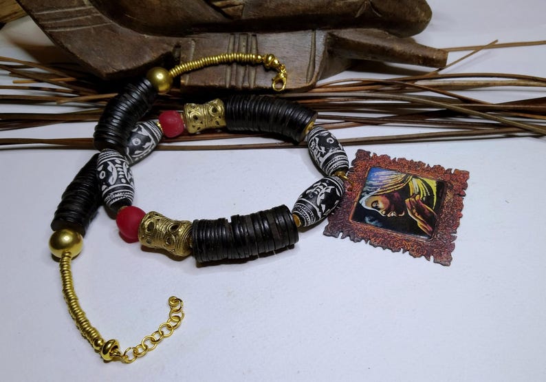 Peut inclure: Un collier artisanal avec des perles noires, dorées, rouges et blanches. Le collier comprend des perles noires cylindriques, des accents métalliques dorés et des perles blanches à motifs. Une petite image encadrée est également présente.