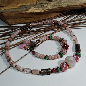 May include: A handmade necklace and bracelet set. The necklace and bracelet feature pink, white, and red beads, with green accents. The necklace has a variety of bead shapes and sizes, including a large white bead with red polka dots.