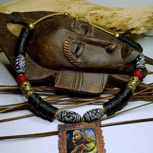May include: A handcrafted necklace featuring black, white, and red beads, with a pendant depicting a woman's portrait. The necklace includes cylindrical and patterned beads, accented with gold-colored metal elements. The pendant is framed in a rustic, textured border.