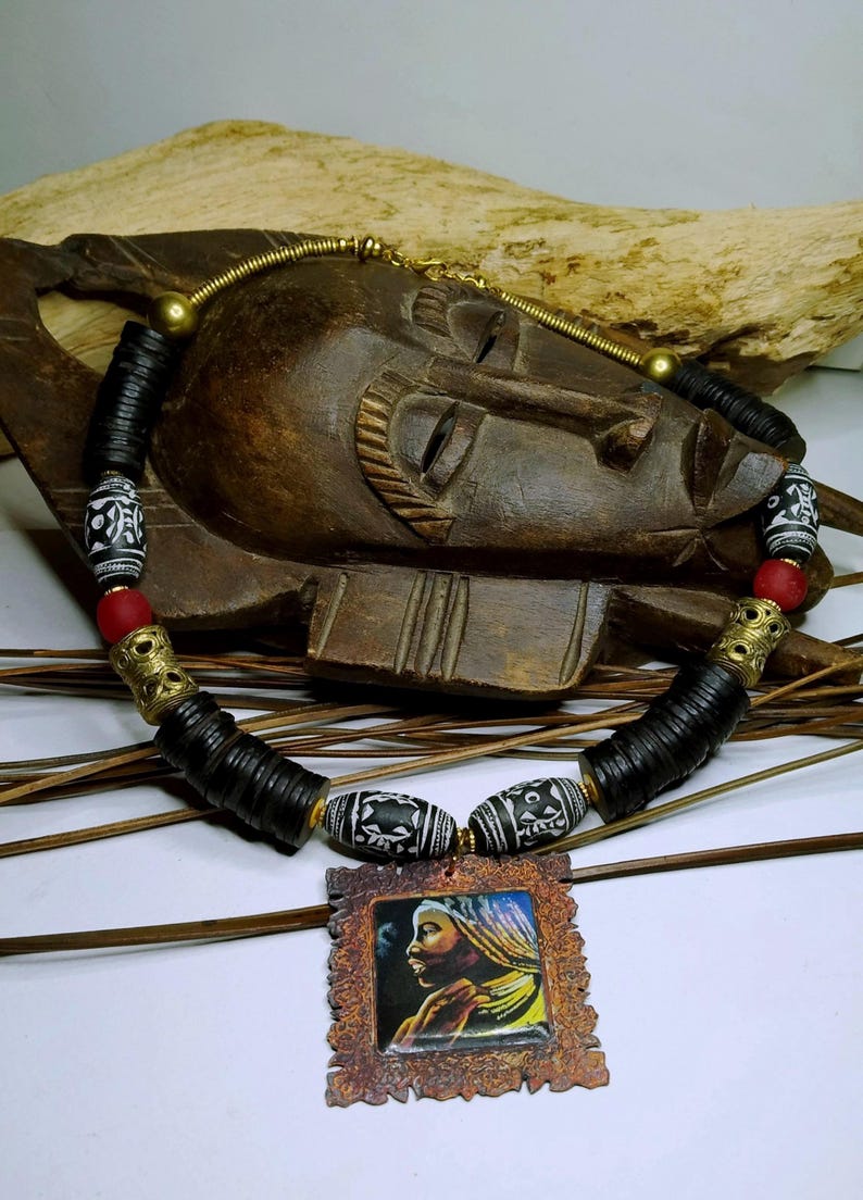 May include: A handcrafted necklace featuring black, white, and red beads, with a pendant depicting a woman's portrait. The necklace includes cylindrical and patterned beads, accented with gold-colored metal elements. The pendant is framed in a rustic, textured border.