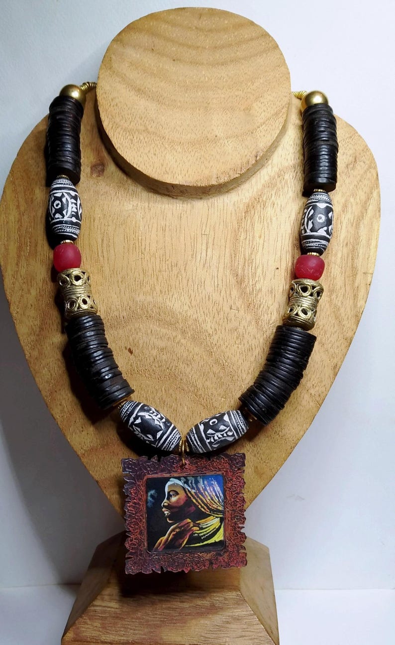 Peut inclure: Collier fait à la main avec des perles en bois noir, des perles blanches et noires à motifs et des perles de verre rouges. Le pendentif est un portrait encadré. Le collier est exposé sur un présentoir en bois.