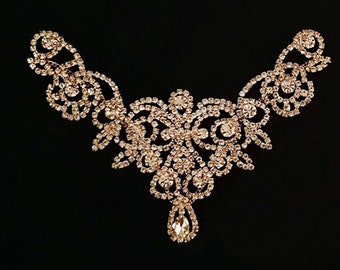 May include: A rose gold-toned necklace with a symmetrical design, featuring intricate scrollwork and numerous clear rhinestones. A teardrop-shaped rhinestone hangs from the center. The necklace is set against a black background.