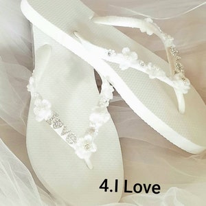 May include: A pair of white flip-flops featuring straps adorned with small white fabric flowers, clear crystals, and silver-toned letters spelling out the word LOVE. The text 4.I Love is printed in black on the white surface of the footwear.