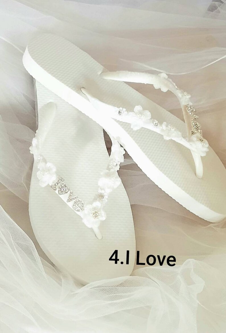 May include: A pair of white flip-flops featuring straps adorned with small white fabric flowers, clear crystals, and silver-toned letters spelling out the word LOVE. The text 4.I Love is printed in black on the white surface of the footwear.