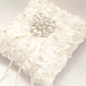 May include: A square white ring pillow covered in fabric rosettes and small faux pearls. A large, circular silver-toned brooch featuring white faux pearls and clear crystals is centered on top, with two long, thin white satin ribbons extending downward.