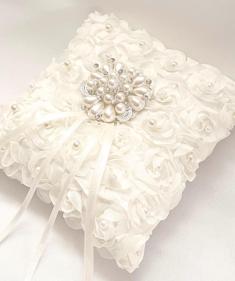 May include: A square white ring pillow covered in fabric rosettes and small faux pearls. A large, circular silver-toned brooch featuring white faux pearls and clear crystals is centered on top, with two long, thin white satin ribbons extending downward.