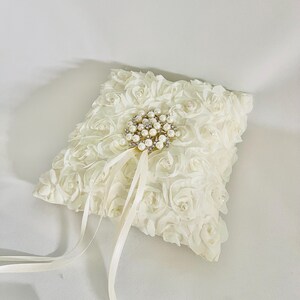 May include: A square off-white ring pillow covered in fabric rosettes with small embedded pearls. A central circular brooch features clusters of white pearls and clear crystals, with two long, thin satin ribbons extending downward from it.