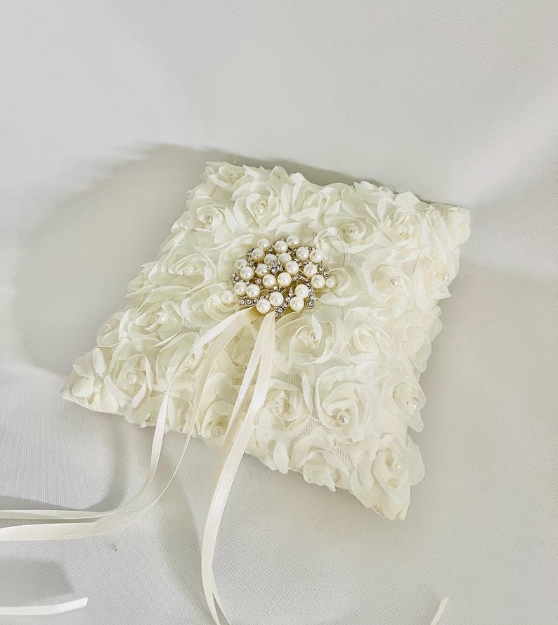 May include: A square off-white ring pillow covered in fabric rosettes with small embedded pearls. A central circular brooch features clusters of white pearls and clear crystals, with two long, thin satin ribbons extending downward from it.