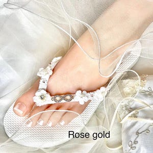 May include: A white flip-flop sandal featuring a textured sole and a strap adorned with white crocheted flowers, small clear crystals, and a central silver-toned infinity symbol. The text "Rose gold" is printed on the lower portion of the image.