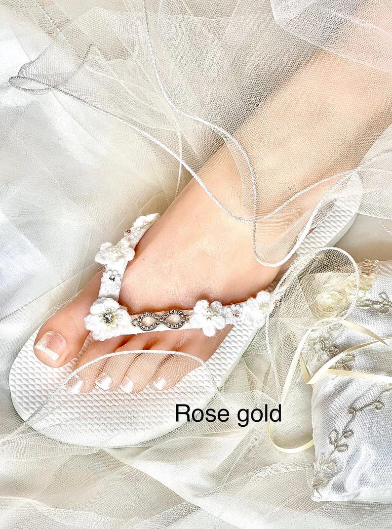 May include: A white flip-flop sandal featuring a textured sole and a strap adorned with white crocheted flowers, small clear crystals, and a central silver-toned infinity symbol. The text "Rose gold" is printed on the lower portion of the image.