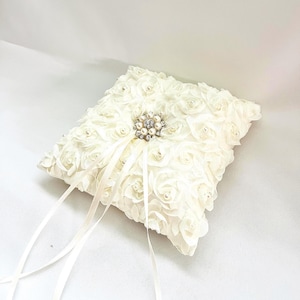 May include: A square off-white ring pillow covered in fabric rosettes and small faux pearls. The center features a decorative brooch made of clear crystals and pearls, with two long, thin satin ribbons extending from the center for tying rings.