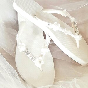 May include: A pair of white flip-flops featuring straps adorned with small white fabric flowers, clear rhinestones, and a silver-toned metal charm that spells out the word love in a cursive script, set against a soft white fabric background.
